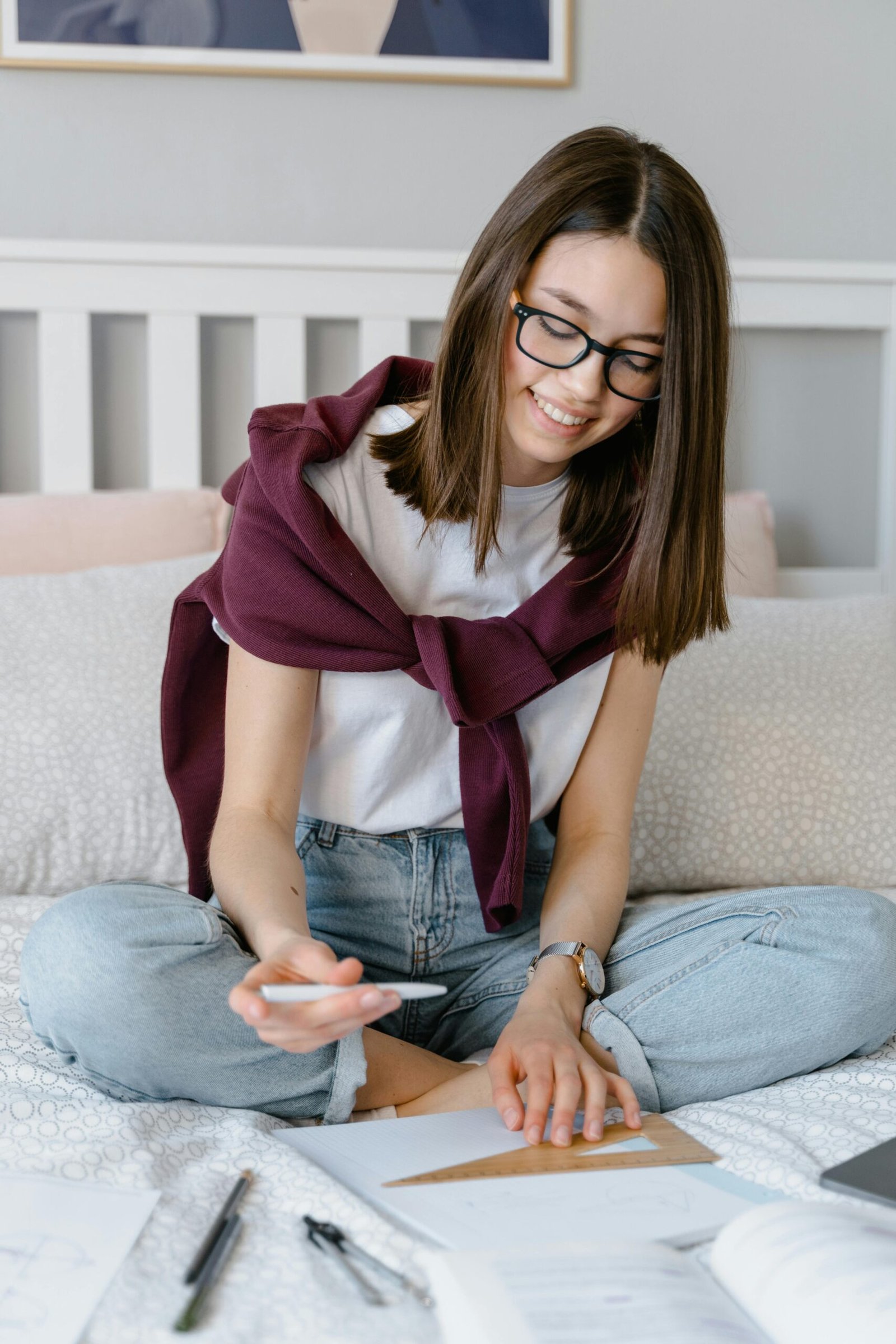 Teen girl with glasses studying on bed, smiling, with notebook and pen.